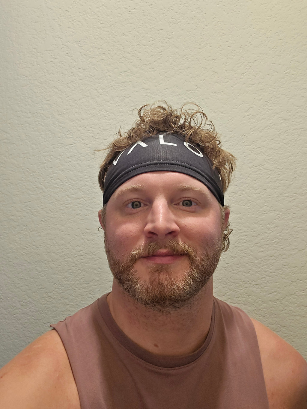 A man wearing a black athletic headband with white VALO lettering and a brown sleeveless shirt, photographed against a plain beige wall.